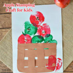 apples stamped on construction paper using a cut apple as the stamp. Brown paper weave basket is placed on the paper below the apples